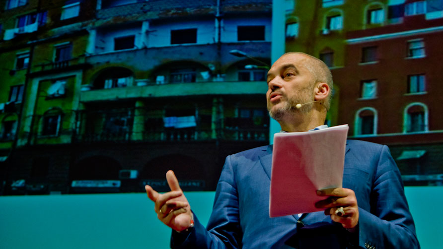 A man gives a presentation in front of projected images of buildings