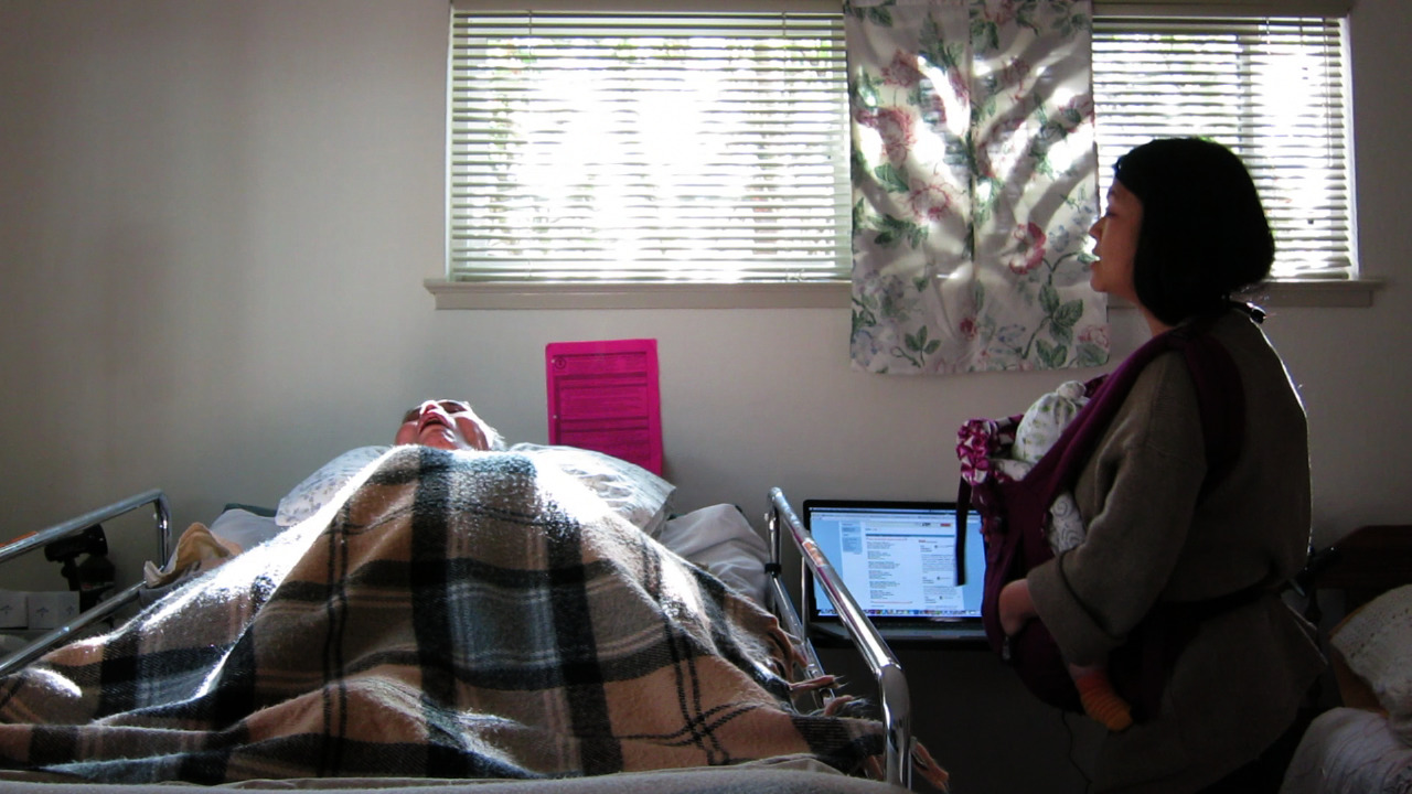 A person holding a baby as they observe someone screaming from a medical bed.