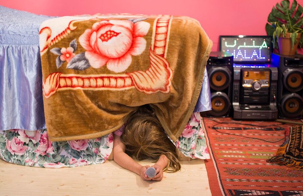 Woman under a bed, pointing a microphone at the camera with a sound system in the background.