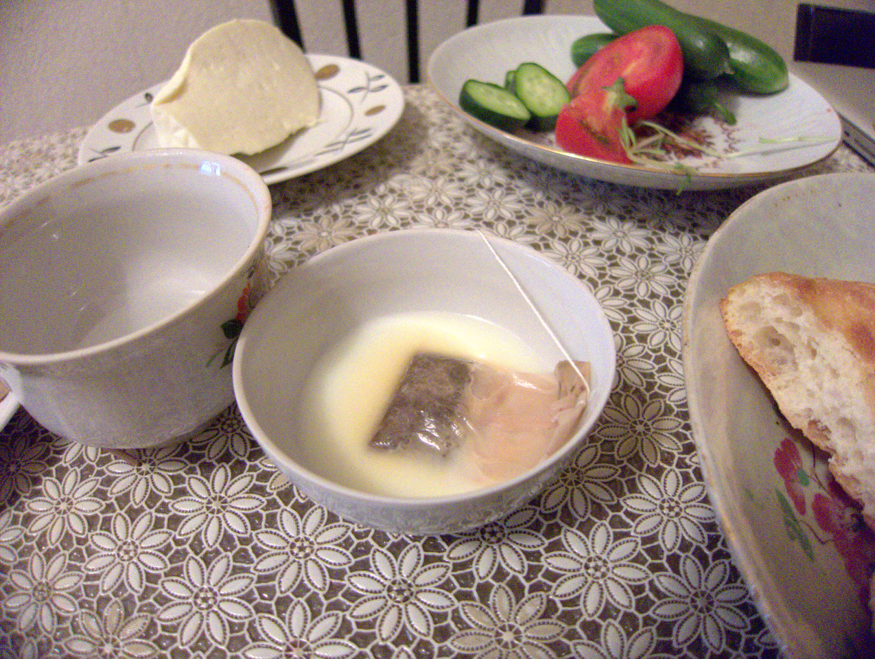 A table with bread,cheese,tea,tomatoes,and cucumbers on different dishes.