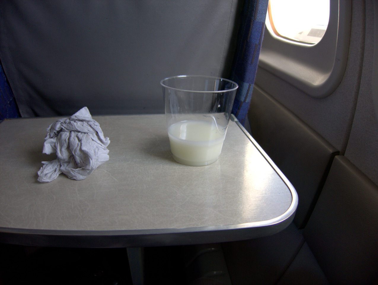 A cup of milk sitting next to a crumbled piece of paper towel on an airplane table.