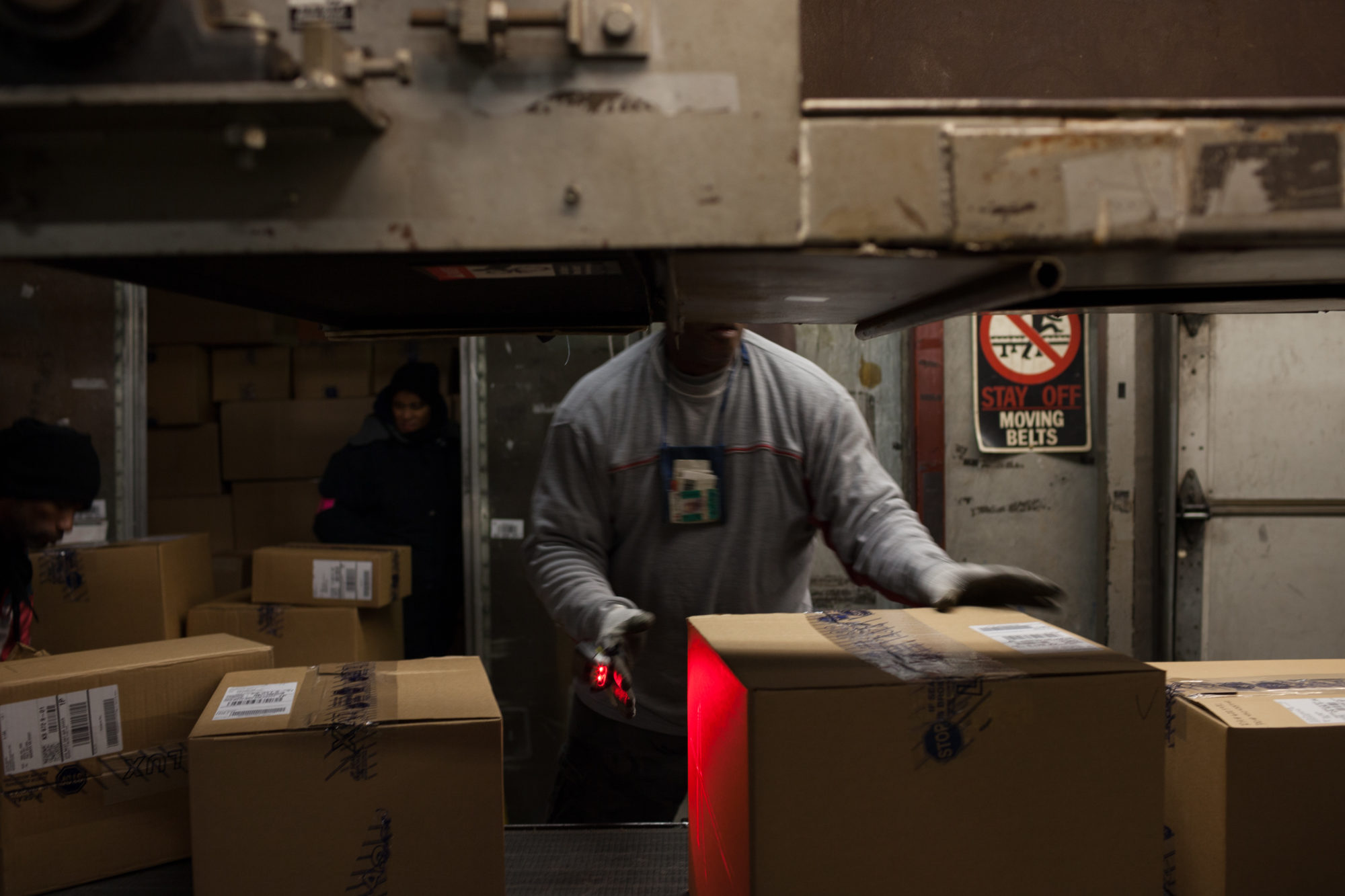 A worker wearing all grey picks a box from the conveyor belt with a red highlight casted on it.