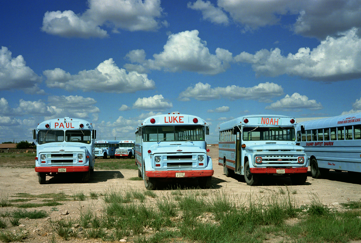 6 blue buses with names on the front in a deserted field