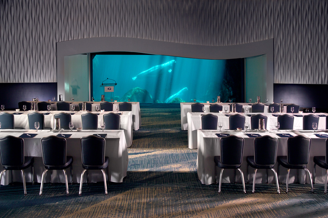 a ballroom with set tables and chairs face a podium with a glass wall behind, showing the aquarium