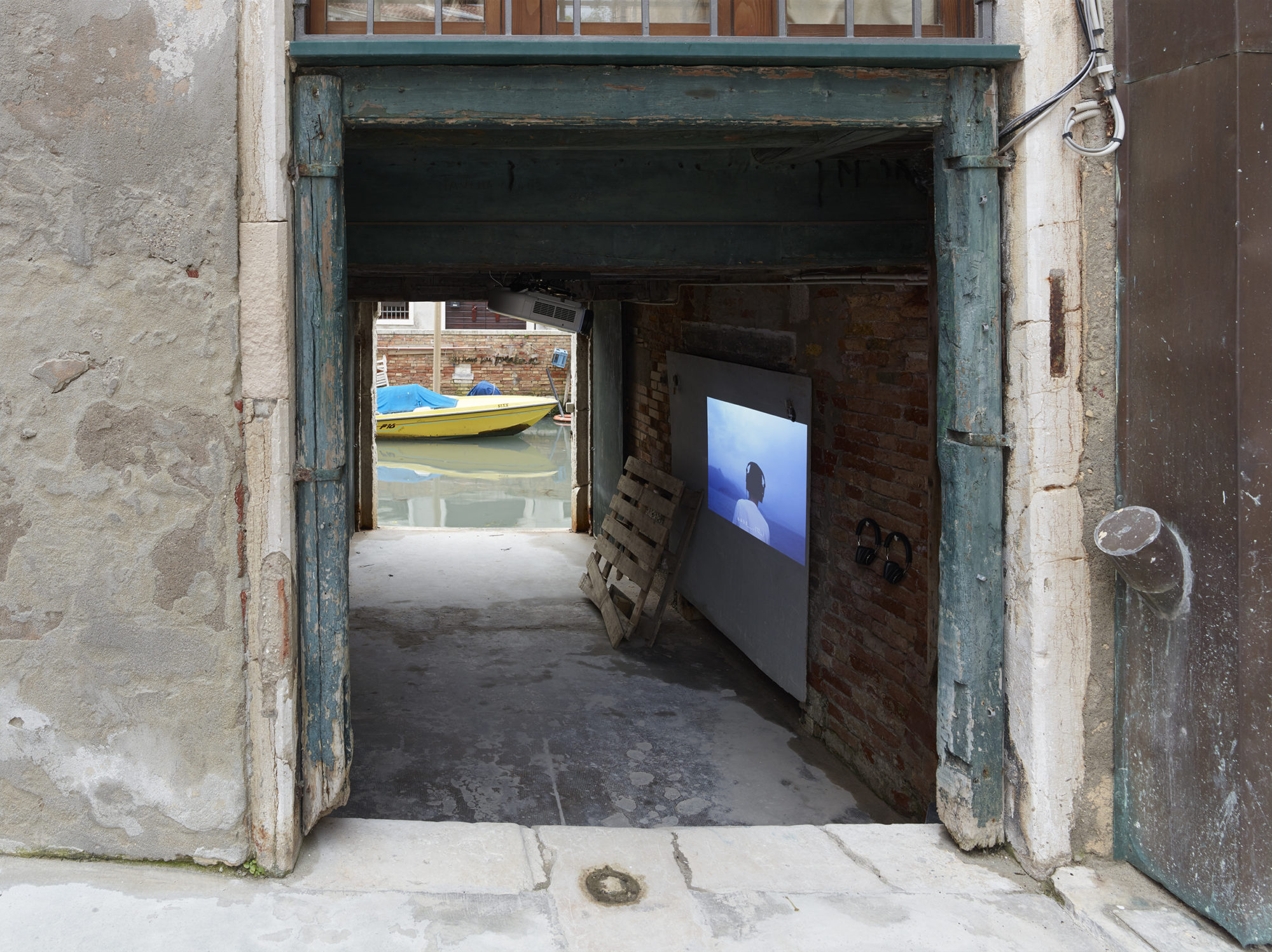 A still from Samson Young's Lullaby (World Music) displayed via projector in an alleyway with a yellow boat floating in the background
