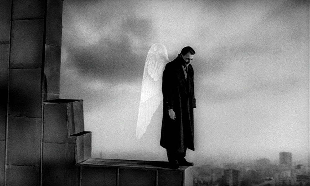 A man with angel wings and a long coat stands on top of a tall building