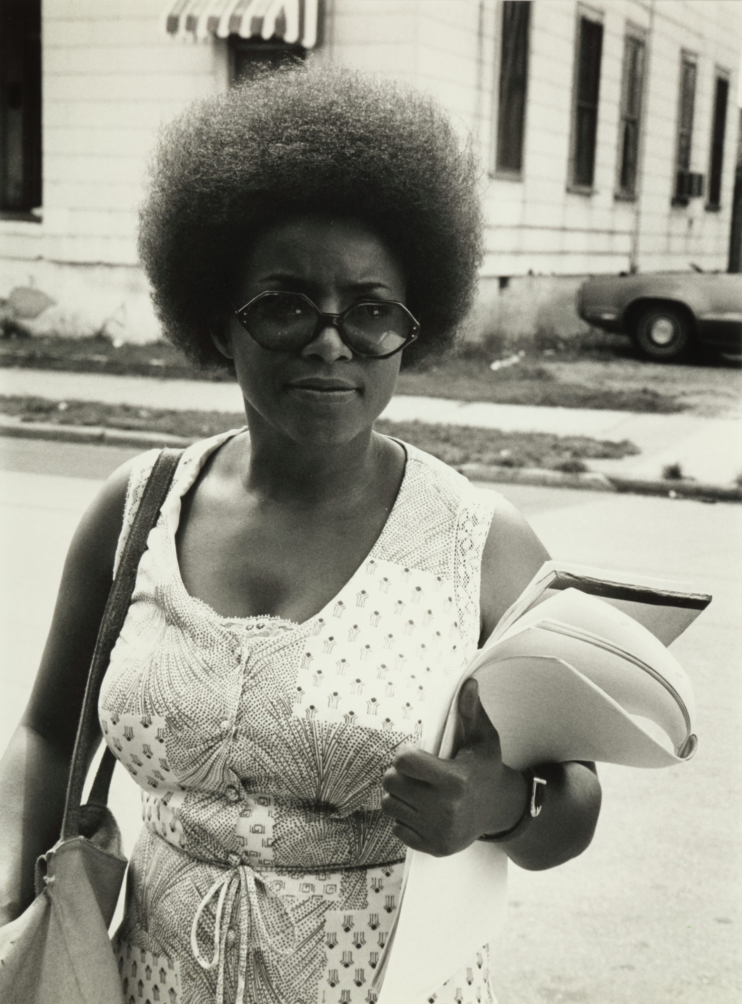 A black and white photograph of a civil rights attorney who is a Black woman with an Afro looking right at the camera and carrying papers in her arms.