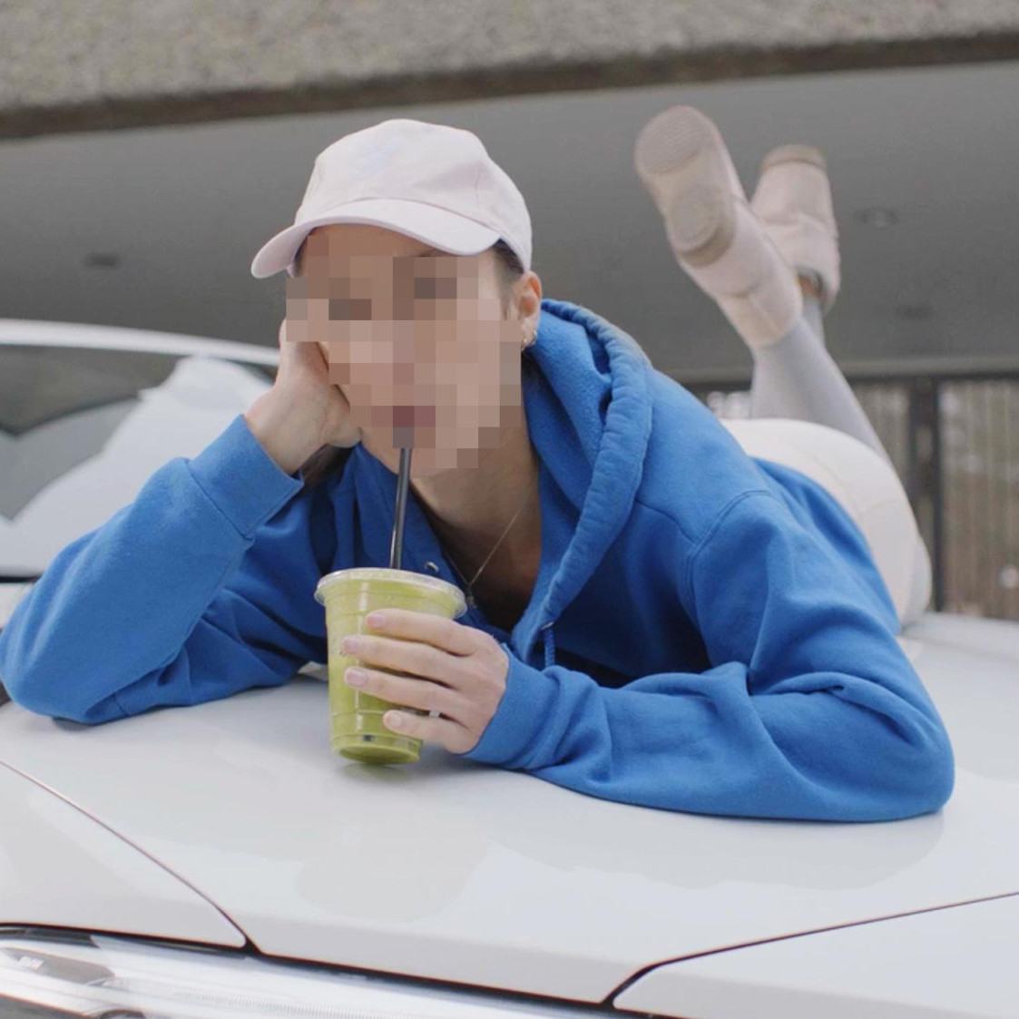 Person in blue hoodie, with face blurred, lays on the hood of a white BMW drinking a green smoothie