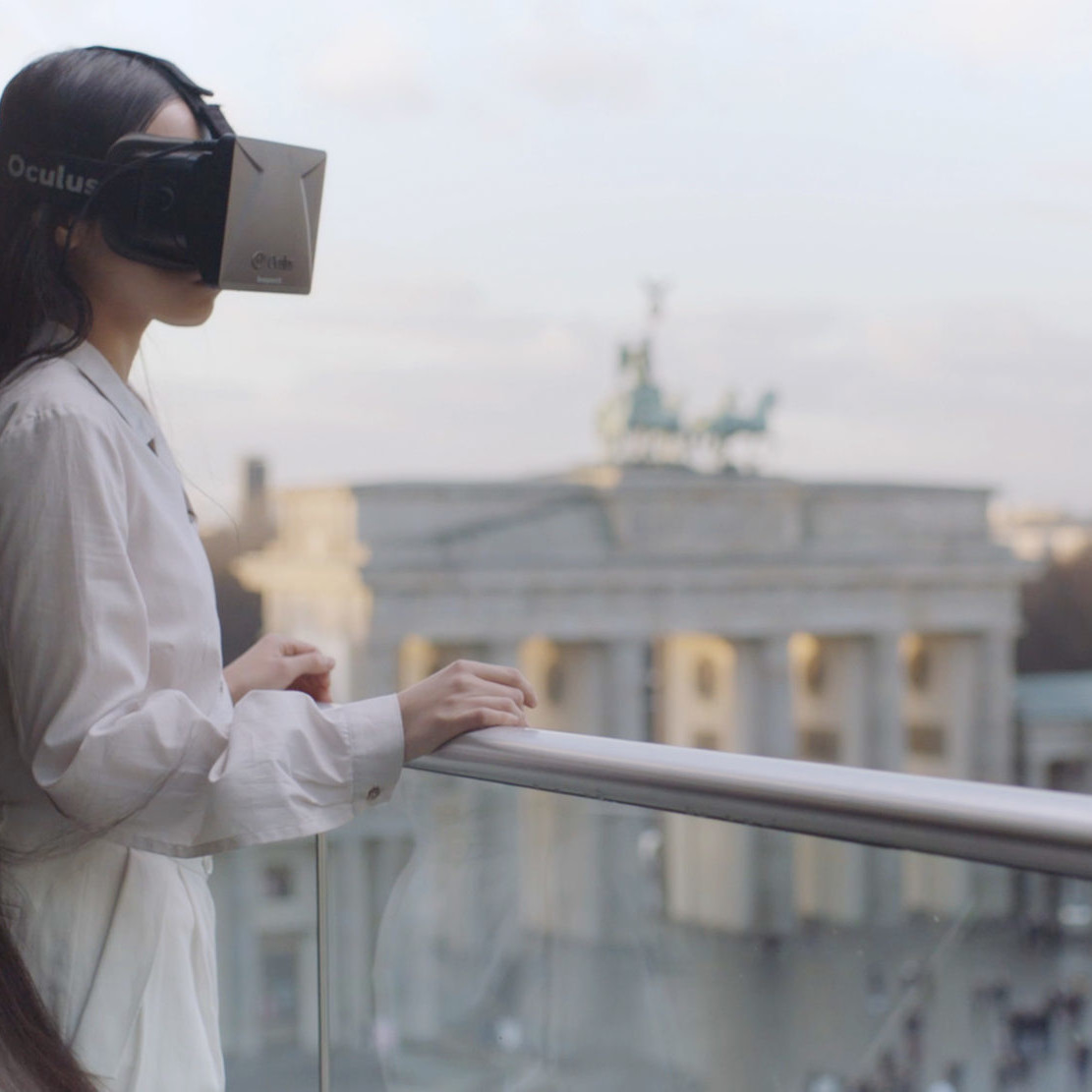 Long-haired woman wearing an Oculus VR headset stands at balcony railing overlooking Pariser Platz in Berlin