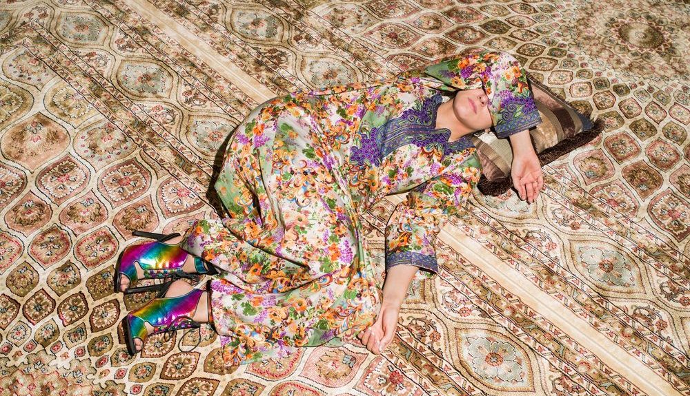 A woman lying on a giant rug wearing a colorful kaftan and rainbow colored heels.