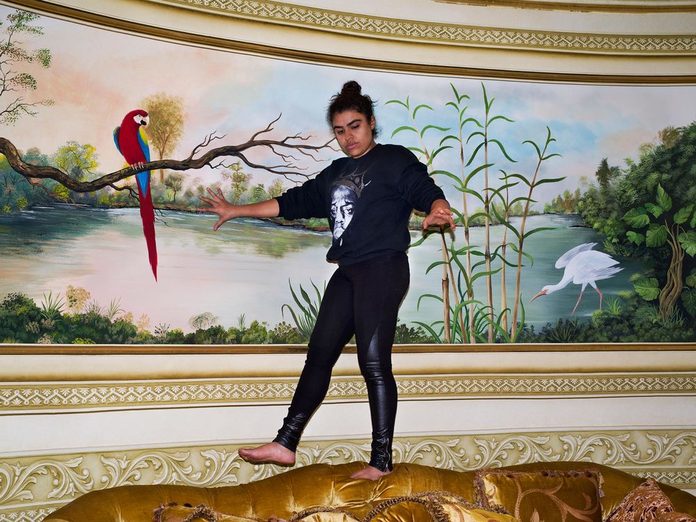 A woman balancing on a vintage couch that's right in front of a tableau featuring a Macaw and a Crane.