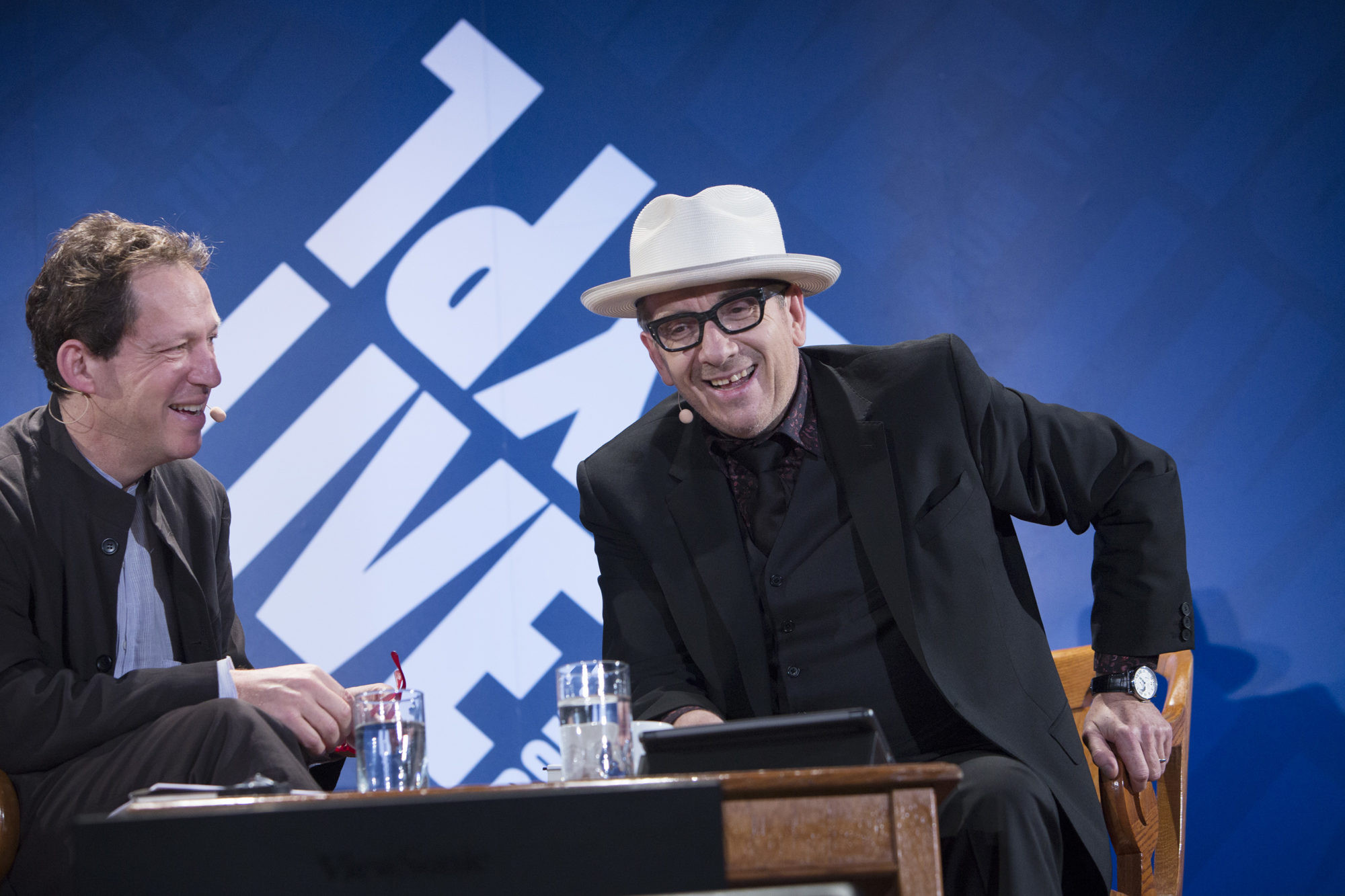 Paul Holdengräber sitting on stage alongside Elvis Costello as they engage in laughter