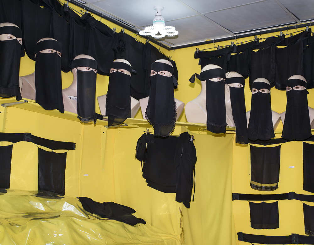 Mannequin busts model black face veils with other veils on hangers behind. Bright yellow fabric covers the walls.