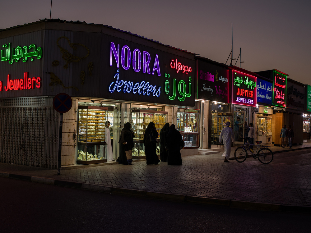 Groups of people walk down a street of storefronts with neon signs at night.