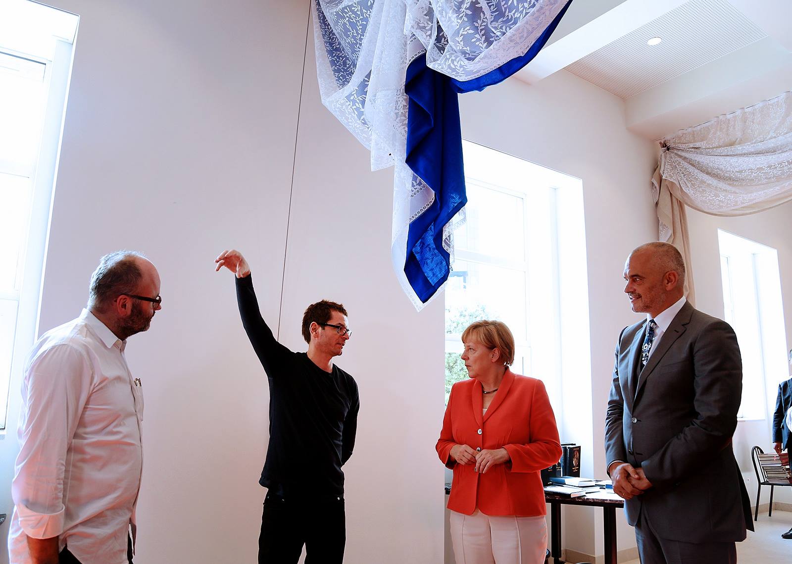 A woman and three men stand and talk in a white room with lace curtains hanging above