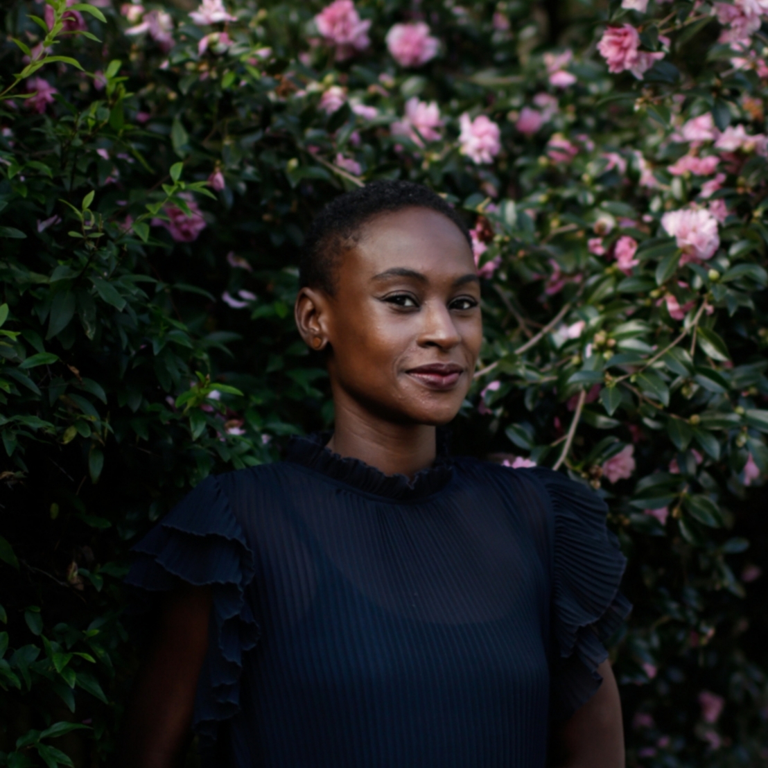 A woman in a dark blue blouse stands against a flowering bush. There is a slight smile on her face.