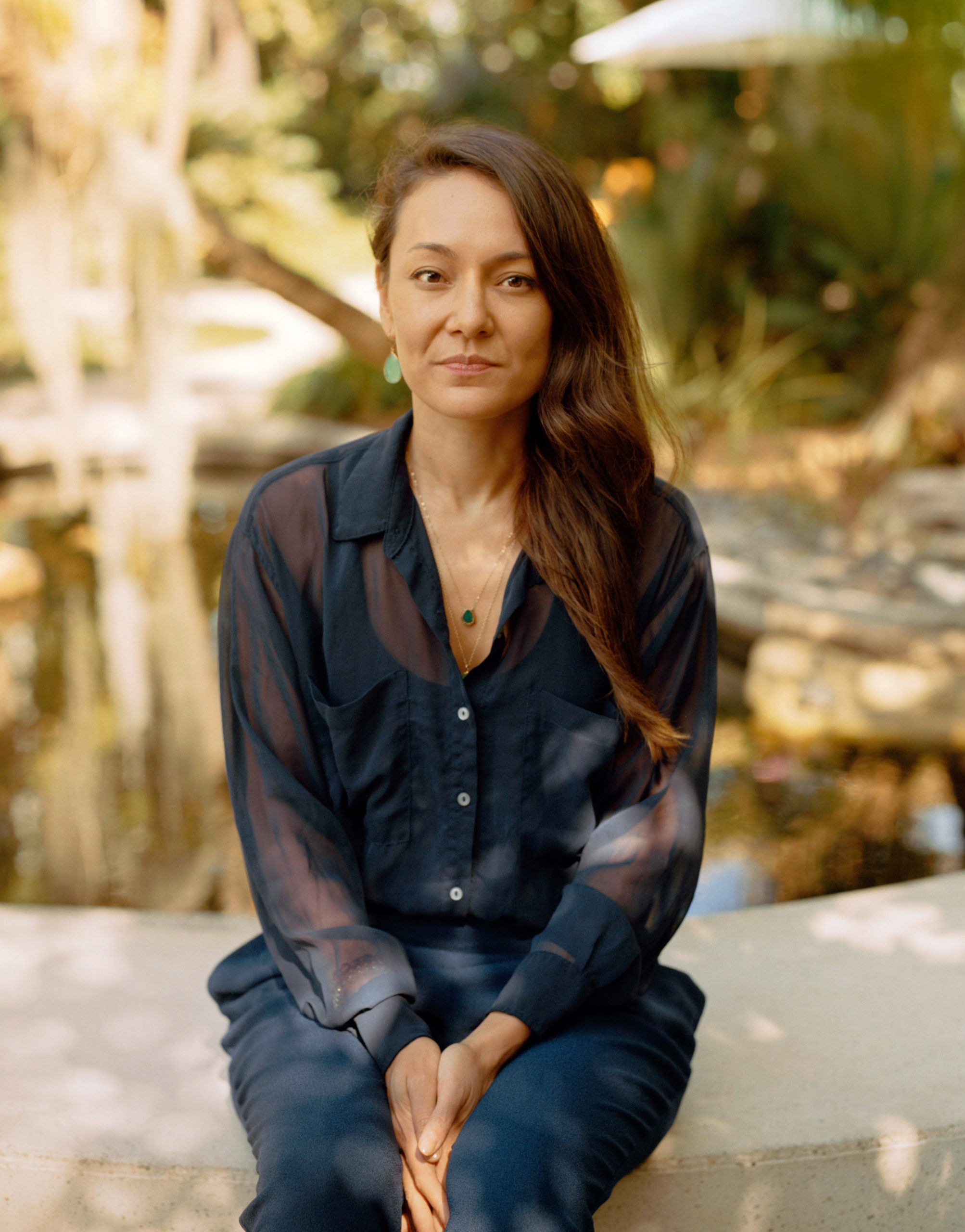 A woman wearing all blue sits on a bench, with a nature scene behind her. Her hair is swept to one side and her hands are placed comfortably between her legs.