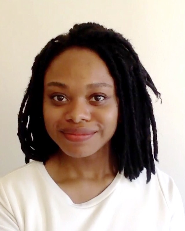 A woman wearing a blouse stands against a white wall. She is smiling.