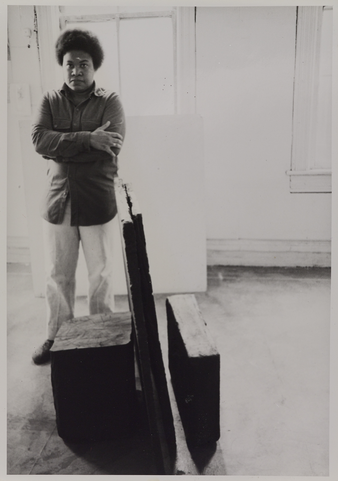 Black and white photograph of Beverly Buchanan, who is standing behind a sculpture with her arms crossed