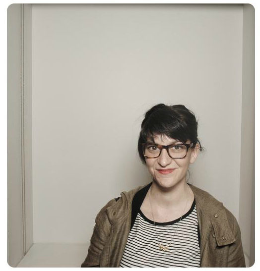 A woman stands in front of a white backdrop. She wears glasses and a striped blouse.