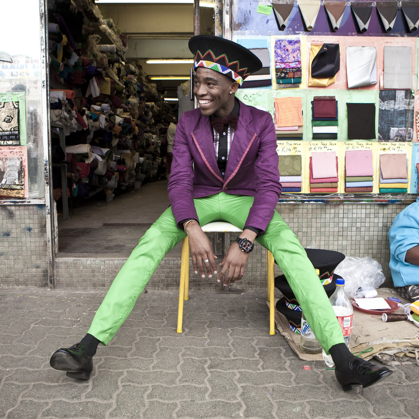 Smiling man wearing a purple blazer, green pants, and black hat sits on a chair in front of a fabric store and a smiling woman in a light blue hoodie