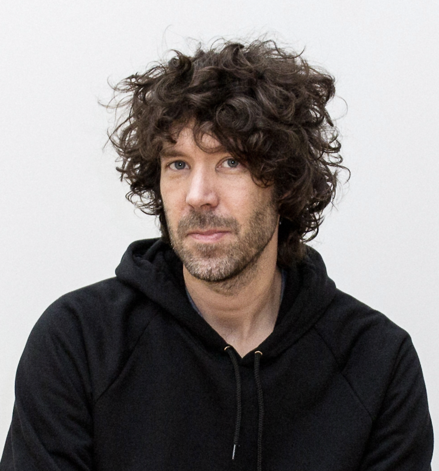 A photograph of a man positioned comfortably in front of a white wall. He wears a black hoodie and has a content expression.