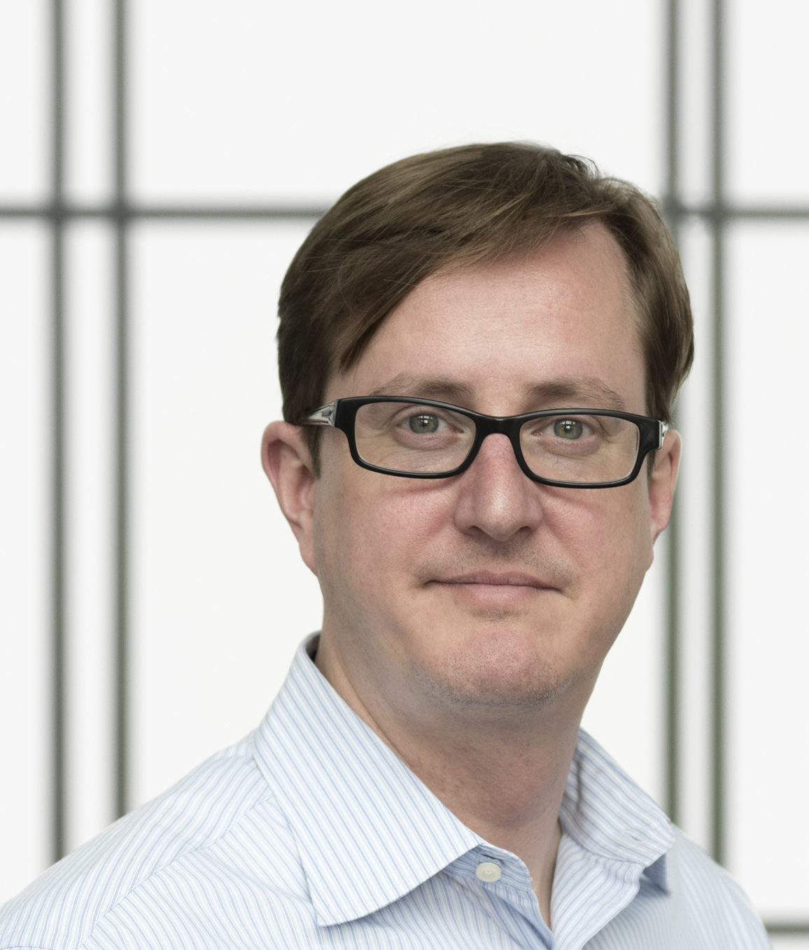 A man wearing a button-up and rectangular glasses appears in front of window panels, gazing at the camera.