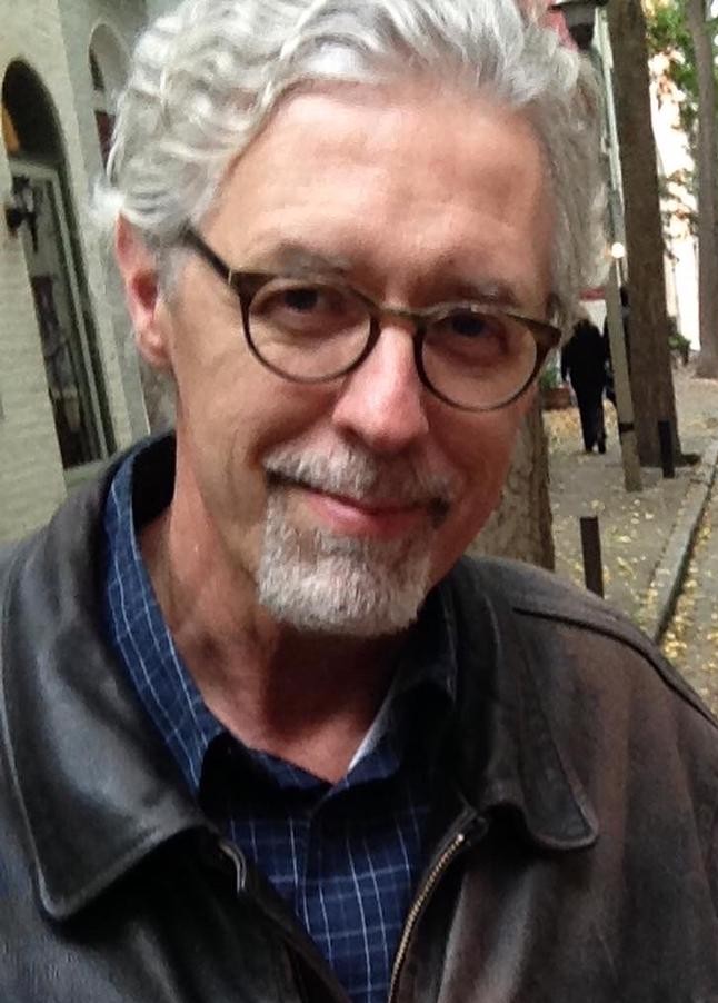 A closely cropped photograph displays a man with glasses. He is wearing a button-up, leather jacket, and round glasses. He appears content.