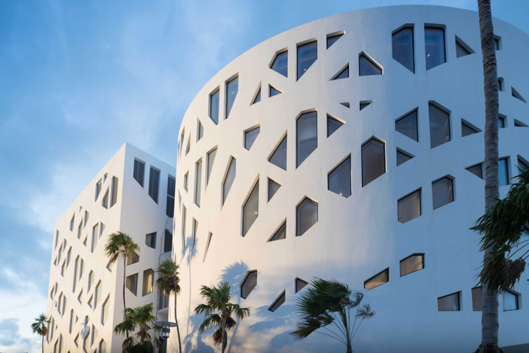 An exterior view of the Faena Forum, featuring a white façade with geometrically irregular windows in a crisscrossing pattern with trees blowing in front of the building.