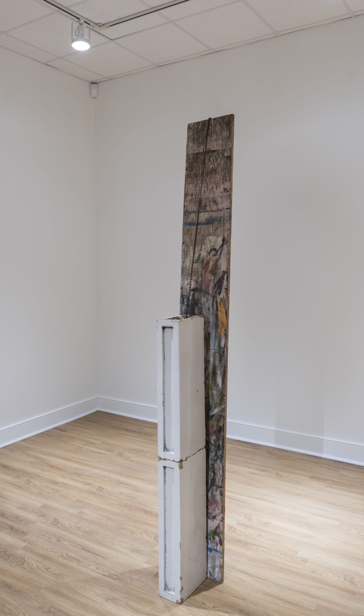A white and brown (with multicolored paint) block-like sculpture by Virginia Overton stands in the corner of a gallery with white floors and ceiling and light-colored wood flooring.