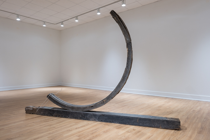 A dark grey sculpture by Virginia Overton, featuring a long plank lying on the floor with a standing semicircle tied to the base, stands in the corner of a gallery with white floors and ceiling and light-colored wood flooring.