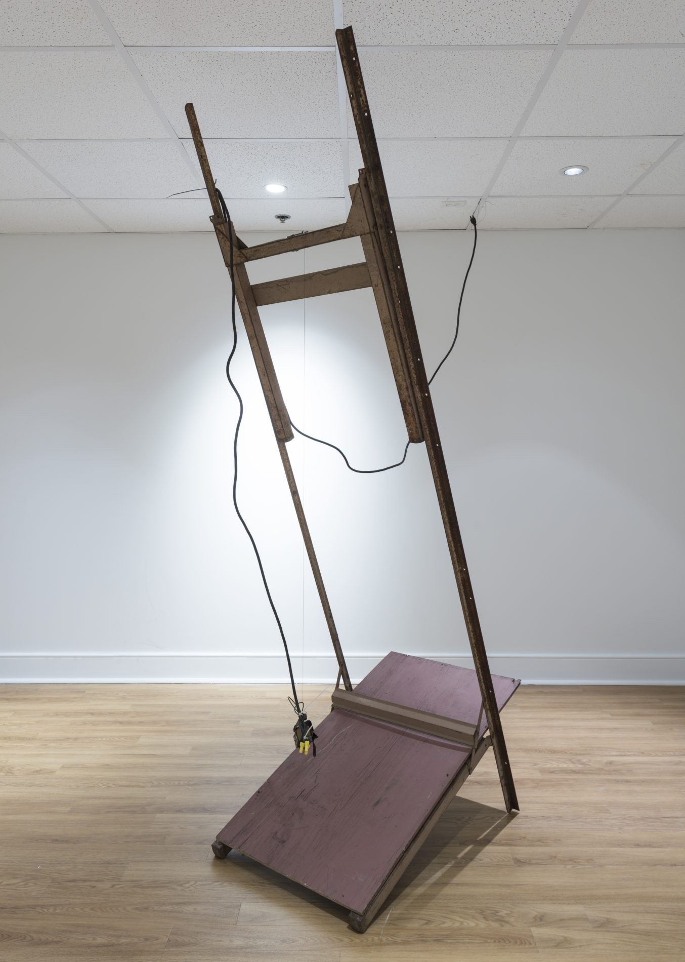 A mostly-wooden sculpture by Virginia Overton leans towards the camera with a black electrical cord hanging off the top of the sculpture. The piece stands in the corner of a gallery with white floors and ceiling and light-colored wood flooring.