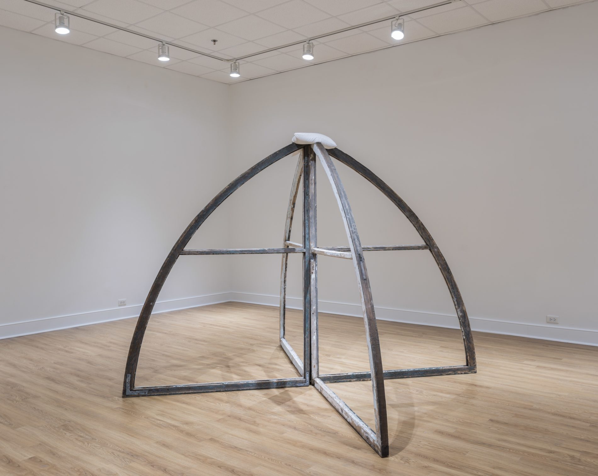 Four-finned dome-like sculpture by Virginia Overton stands in the corner of a gallery with white floors and ceiling and light-colored wood flooring.