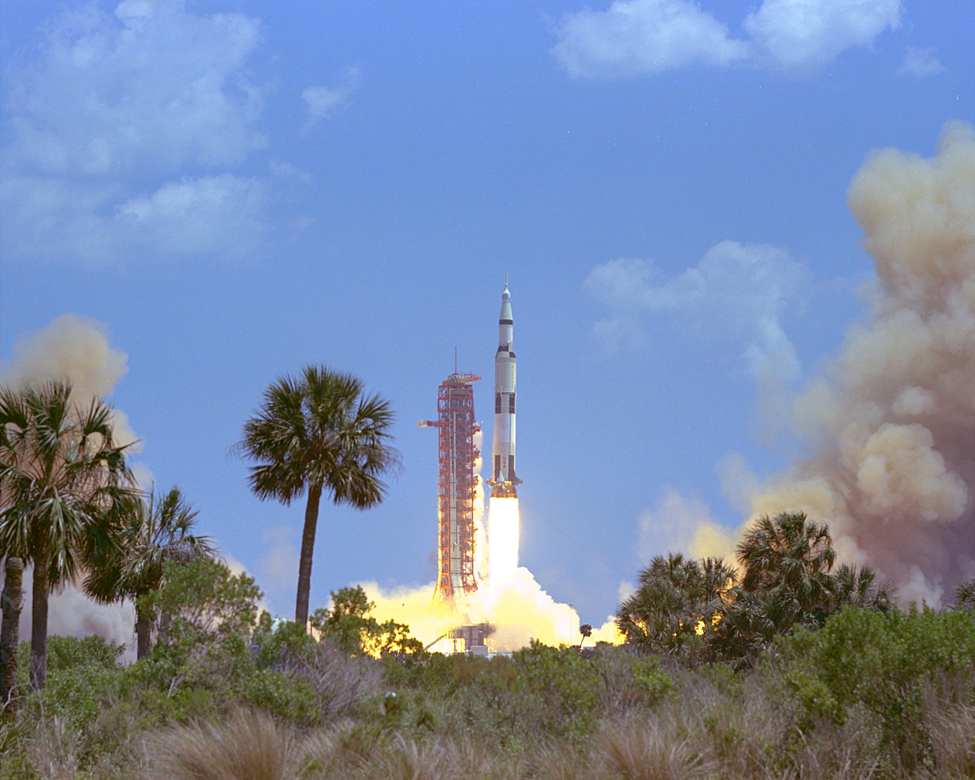 The white and black Apollo 16 rocket ship blasts off from the red launch pad against a blue sky with green and brown palm trees and brush in the foreground