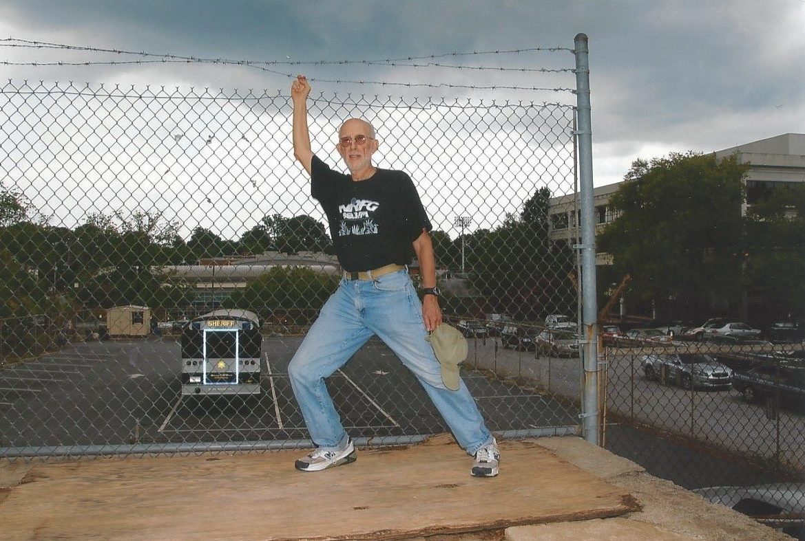 Brian Sherman, wearing blue jeans and a black t shirt, poses in front of a fence with a bus in the background