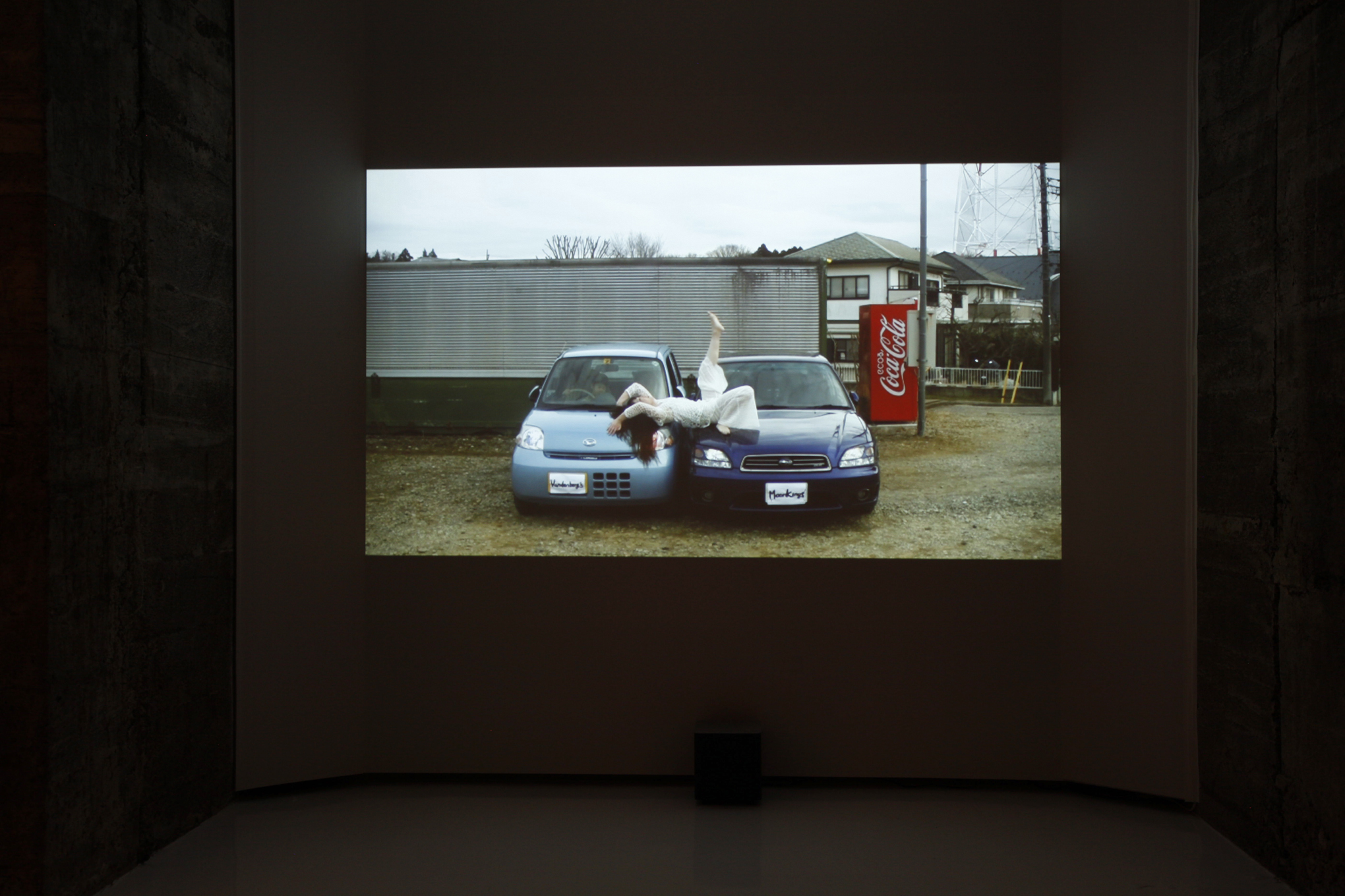 A video still of a woman laying on top of two blue cars while in a yard