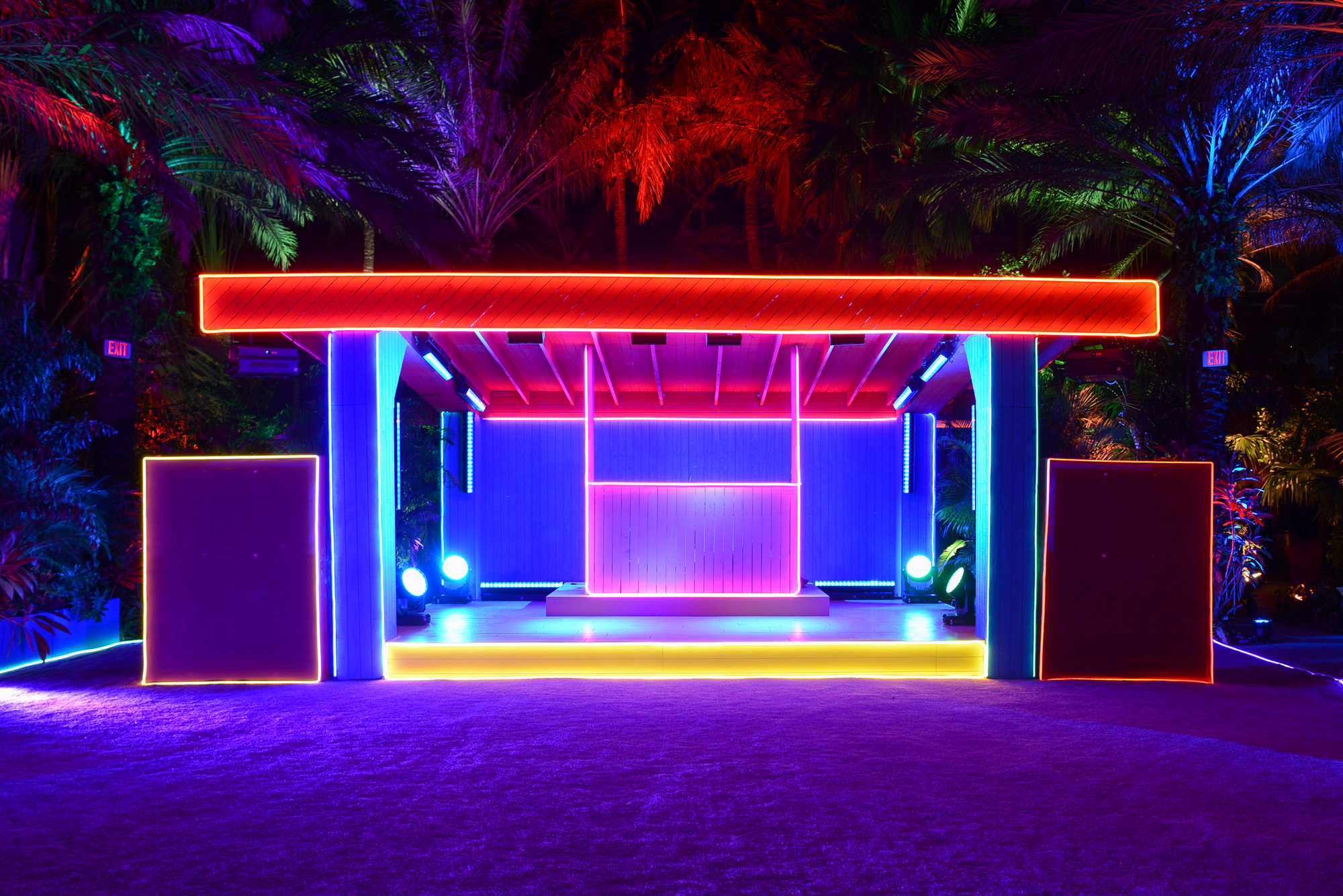 A symmetrical, blocky pavilion serving as part of the Prada Double Club Miami is outlined with lines of neon lights in shades of red, pink, blue, yellow, and orange. The sand in front of the pavilion has a violet hue, and the trees in the background are illuminated by green, blue, red, and purple lights.