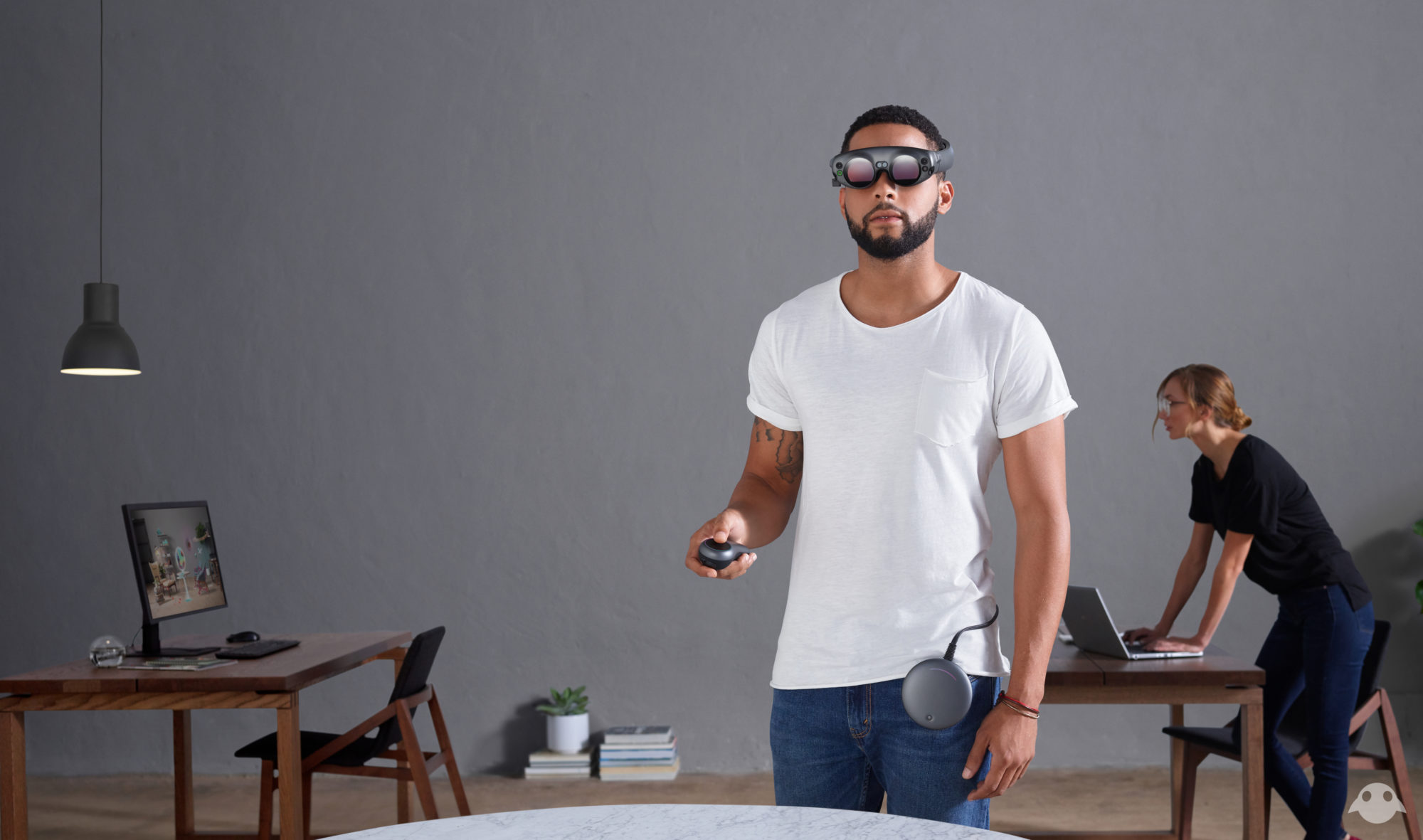 A man wearing a white t-shirt and blue jeans looks into the distance while holding a remote in his hand and wearing a Magic Leap VR headset. In the background, there are two wooden workstations with computers and a woman wearing blue jeans and a black t-shirt standing behind one computer.