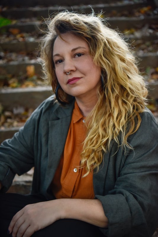 A woman with wavy hair wearing an orange shirt and gray jacket against stairs with leaves on them.