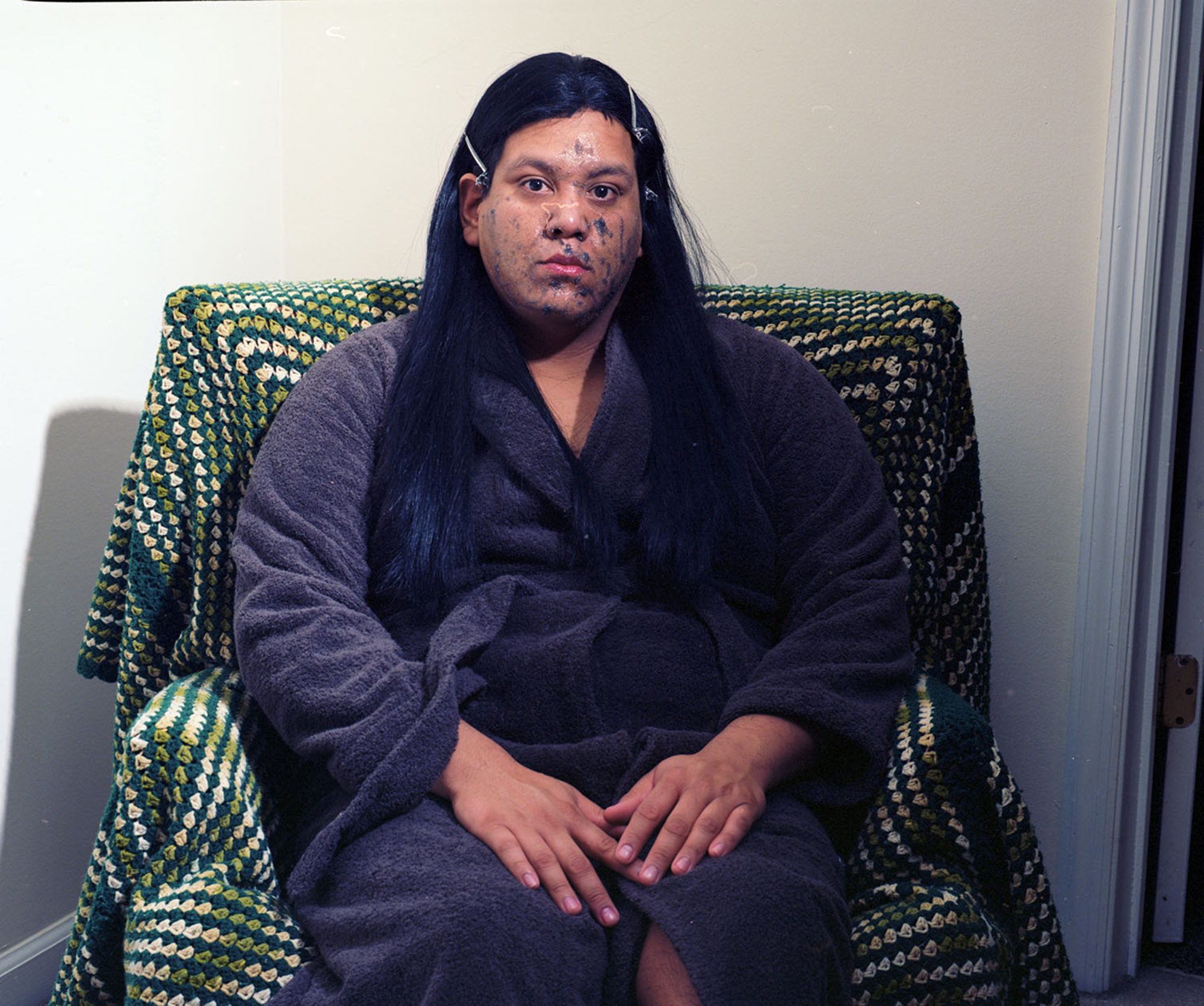 A woman sits in a chair, in a robe, with black makeup smeared on her face.