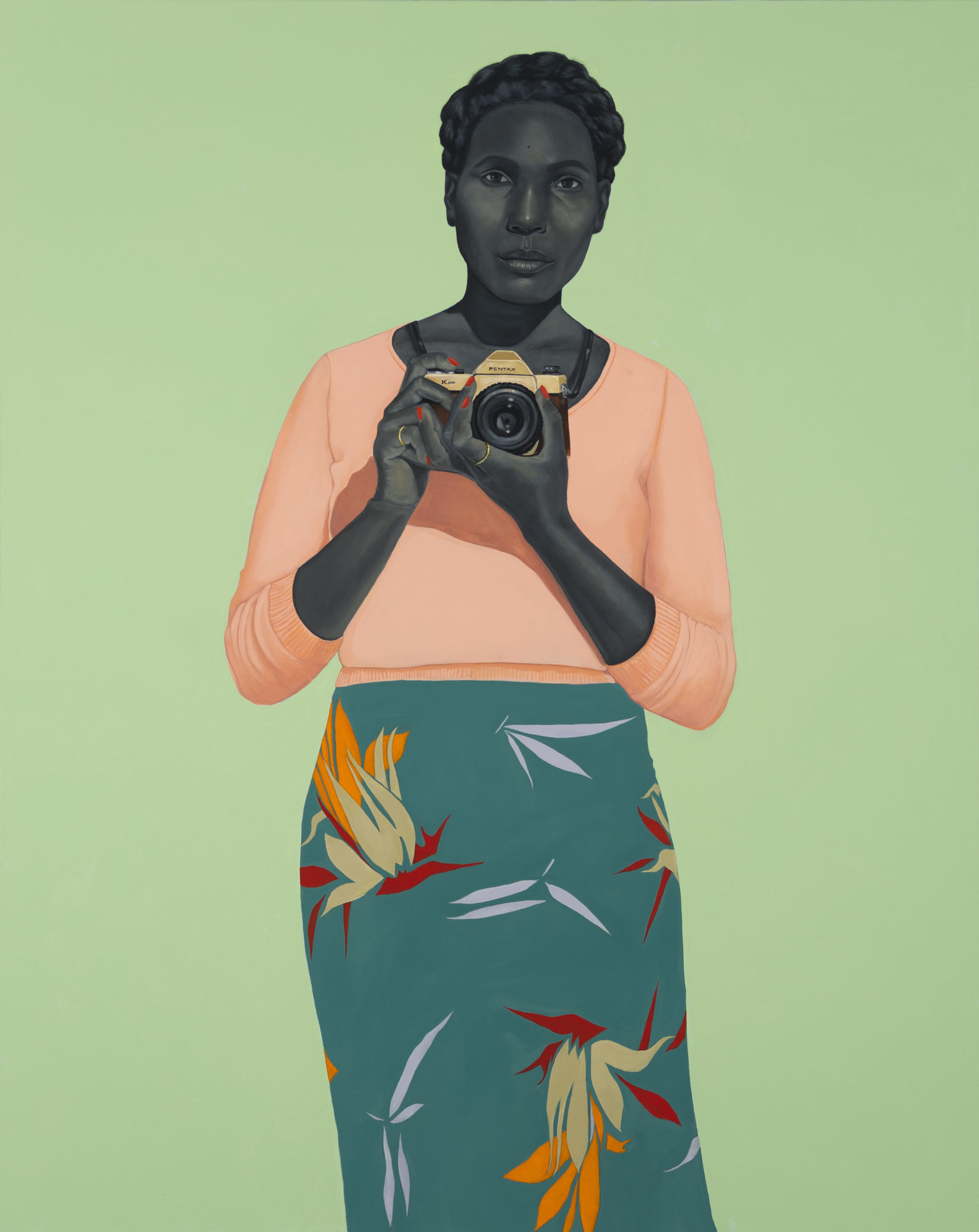 A woman holding a camera, wearing a floral skirt, holds a camera in front of a green background
