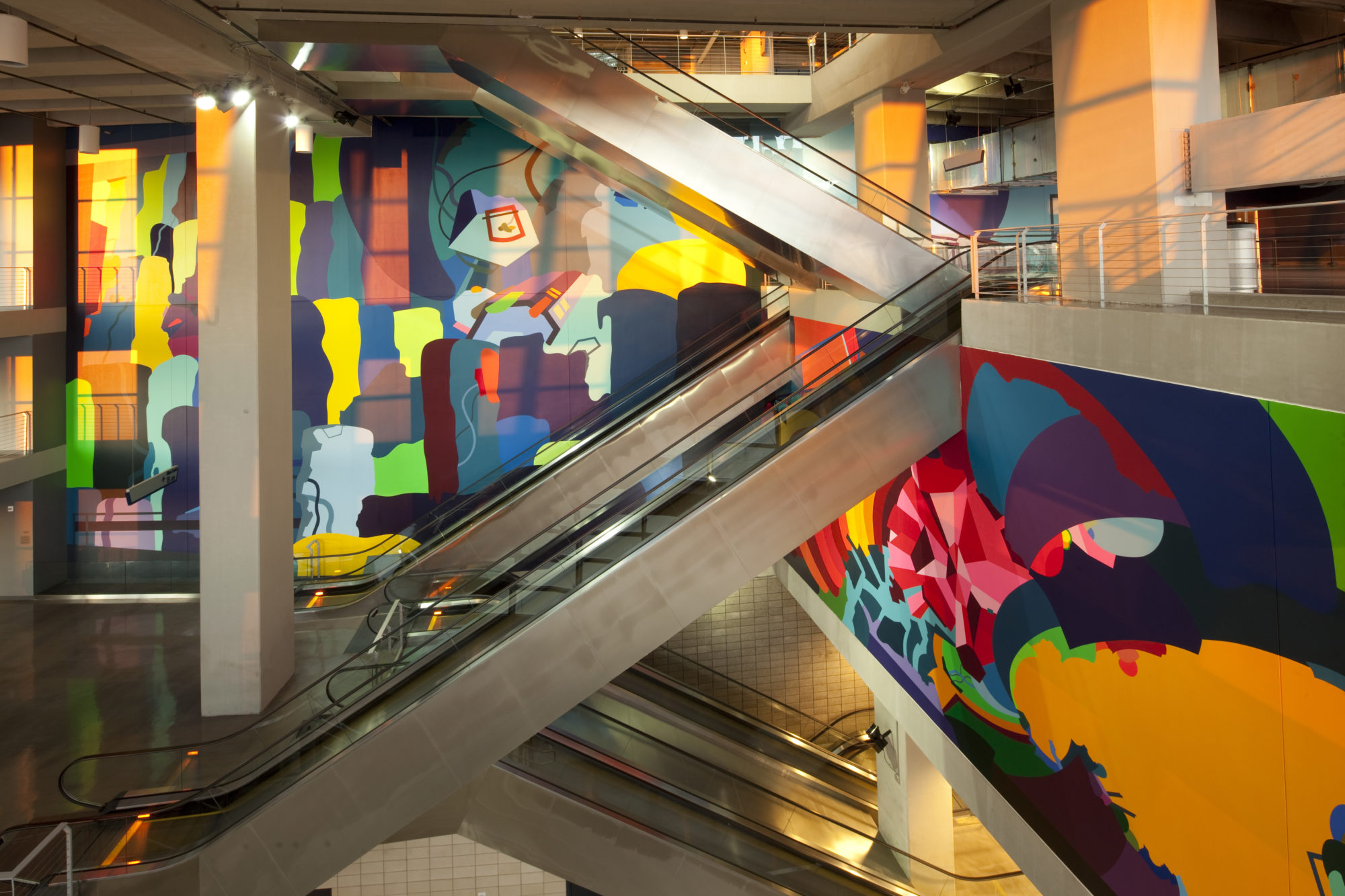 A view of natural light pouring onto an abstract mural made up of purple, yellow, orange, lime green, aqua, navy, cherry red, hot pink, maroon and lilac, covering the Southwest Monumental Staircase of the AT&T stadium.