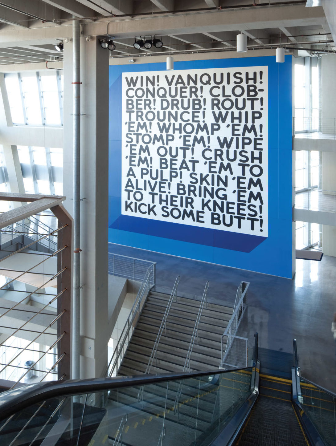 Painted black text appears on a large white wall, with a thick blue border in the Northeast Monumental Staircase of the AT&T stadium. The text reads: 