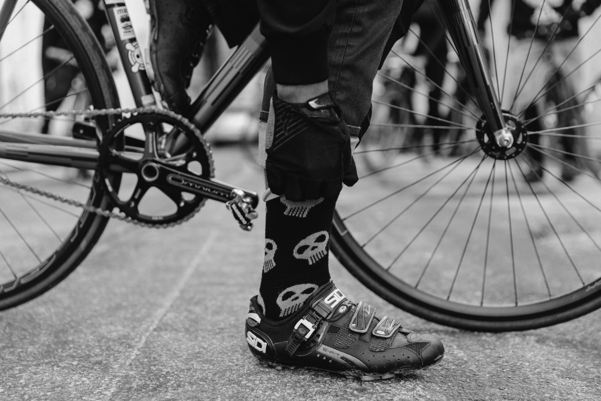 A black and white picture of a foot with bicycle shoes and skull socks propping up a bicycle