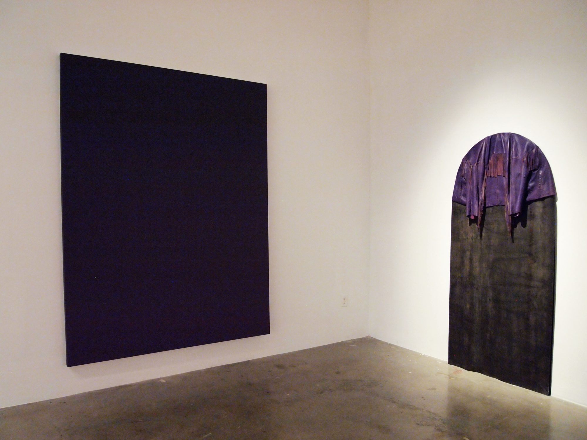 gallery scene showing a large black canvas and a a large, black tombstone shaped piece with a purple leather jacket draped across the top