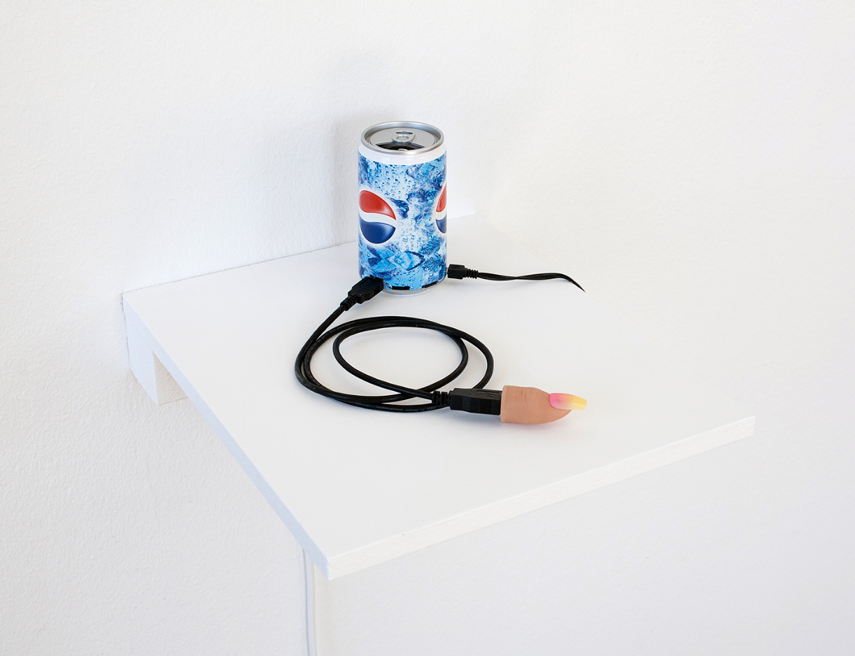 A soda can on a white shelf with two black cables connected to it, one of which is connected to a plastic thumb.