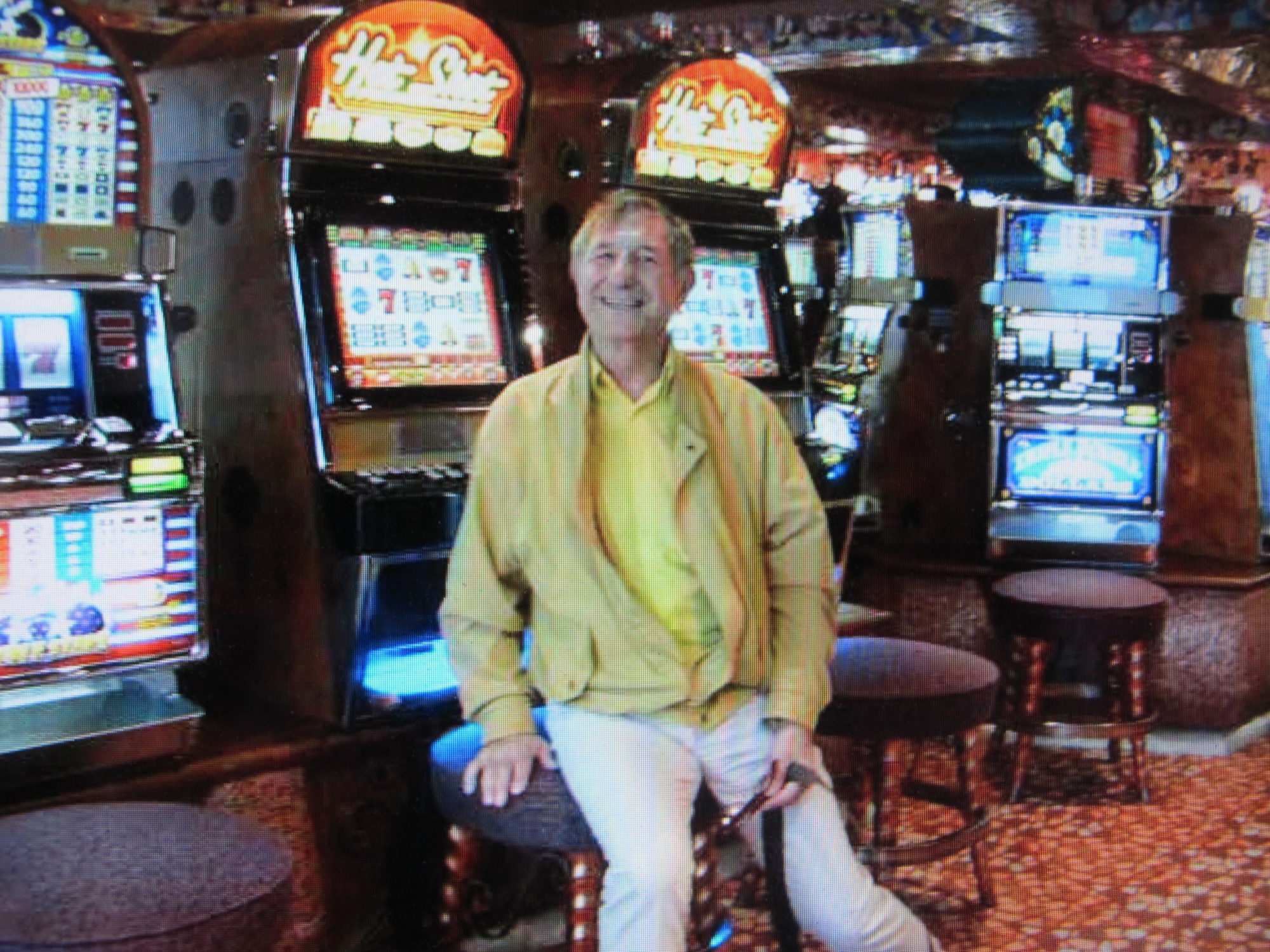 older man in yellow and khaki smiling on a stool in a casino