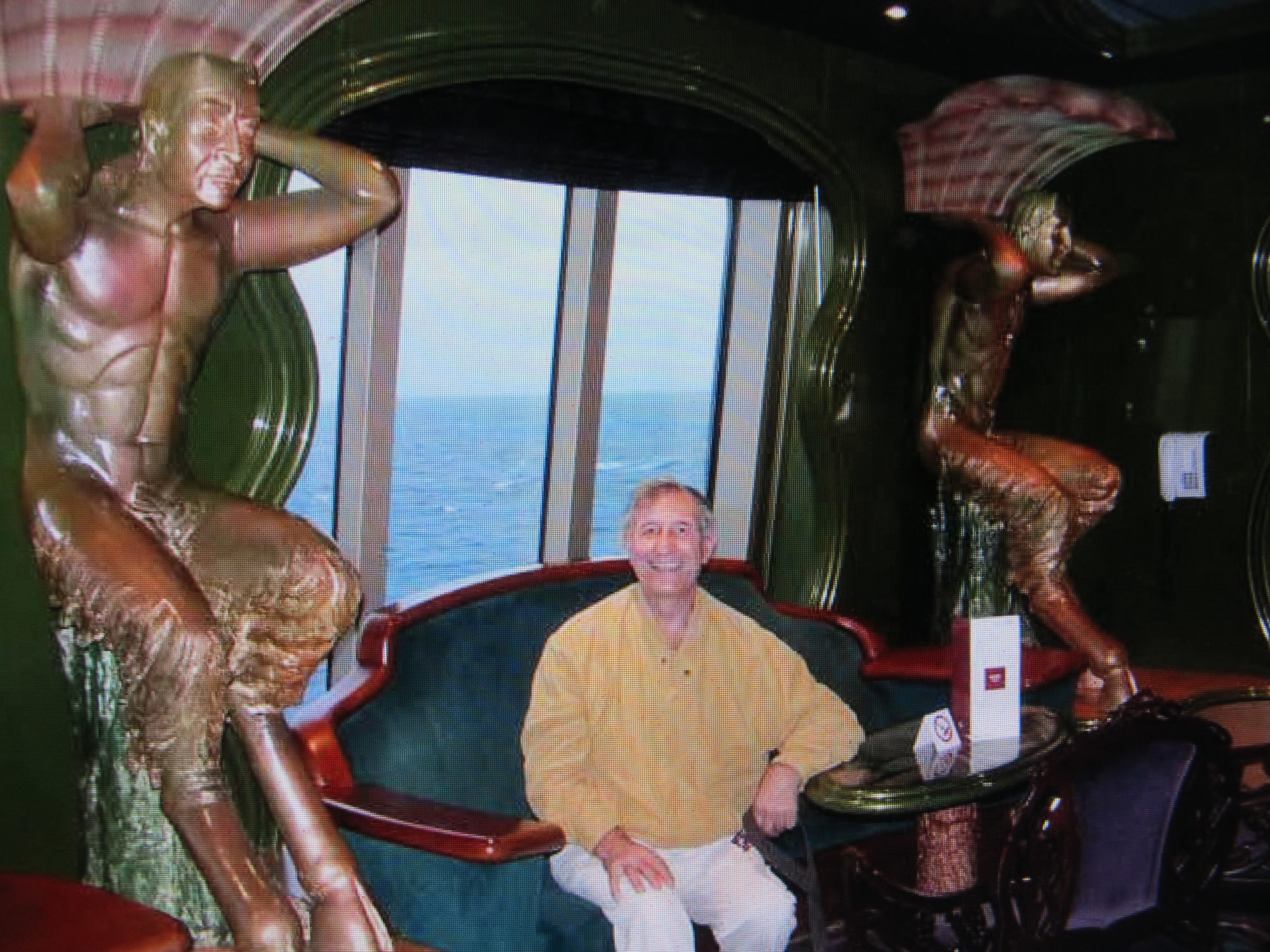 older man in yellow and khaki smiling in a fancy room on a cruise ship showing the ocean