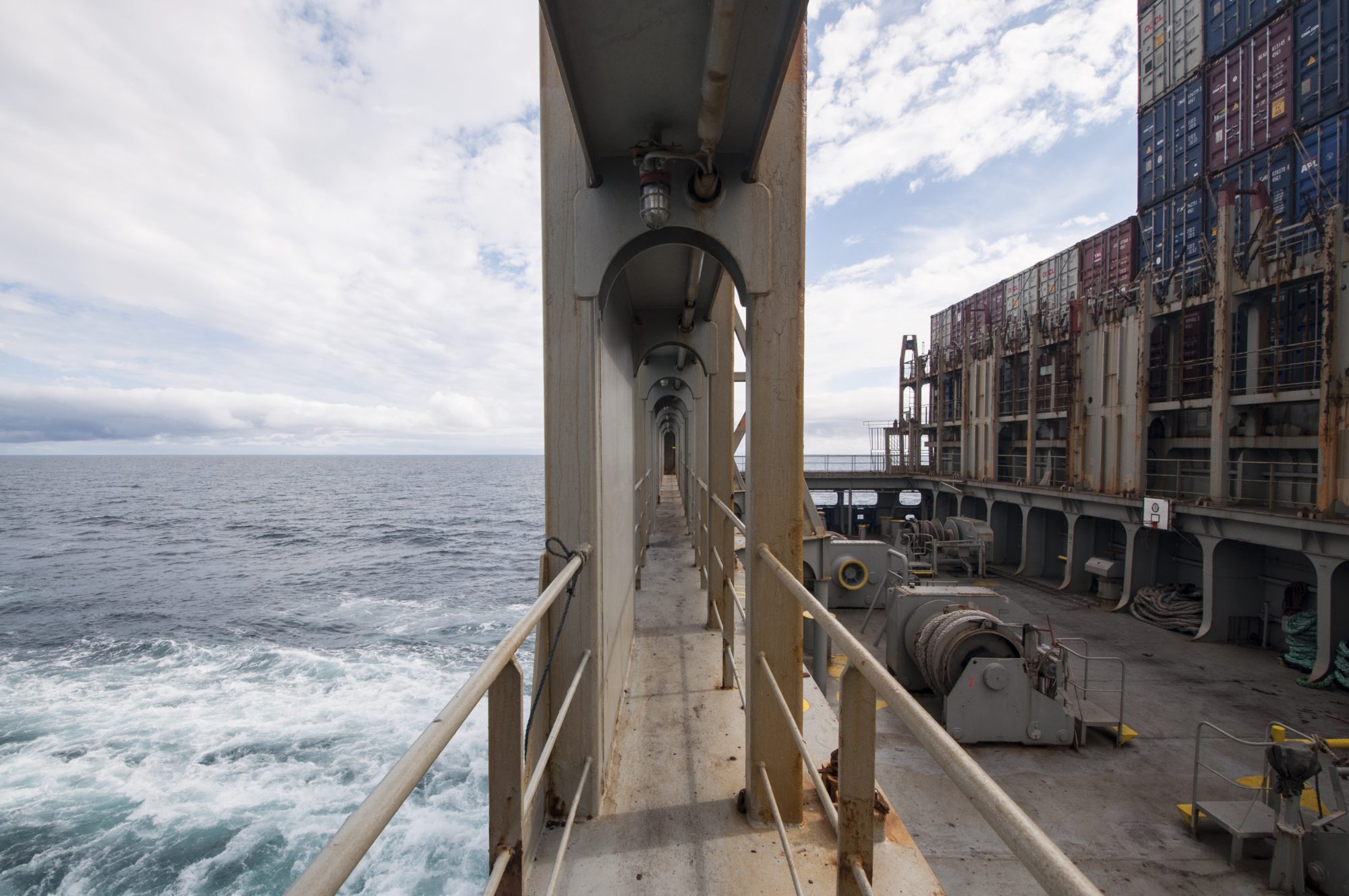 image split in half by cargo ship walkway, vastness of water on the left, shipping containers and cargo derricks on the right.