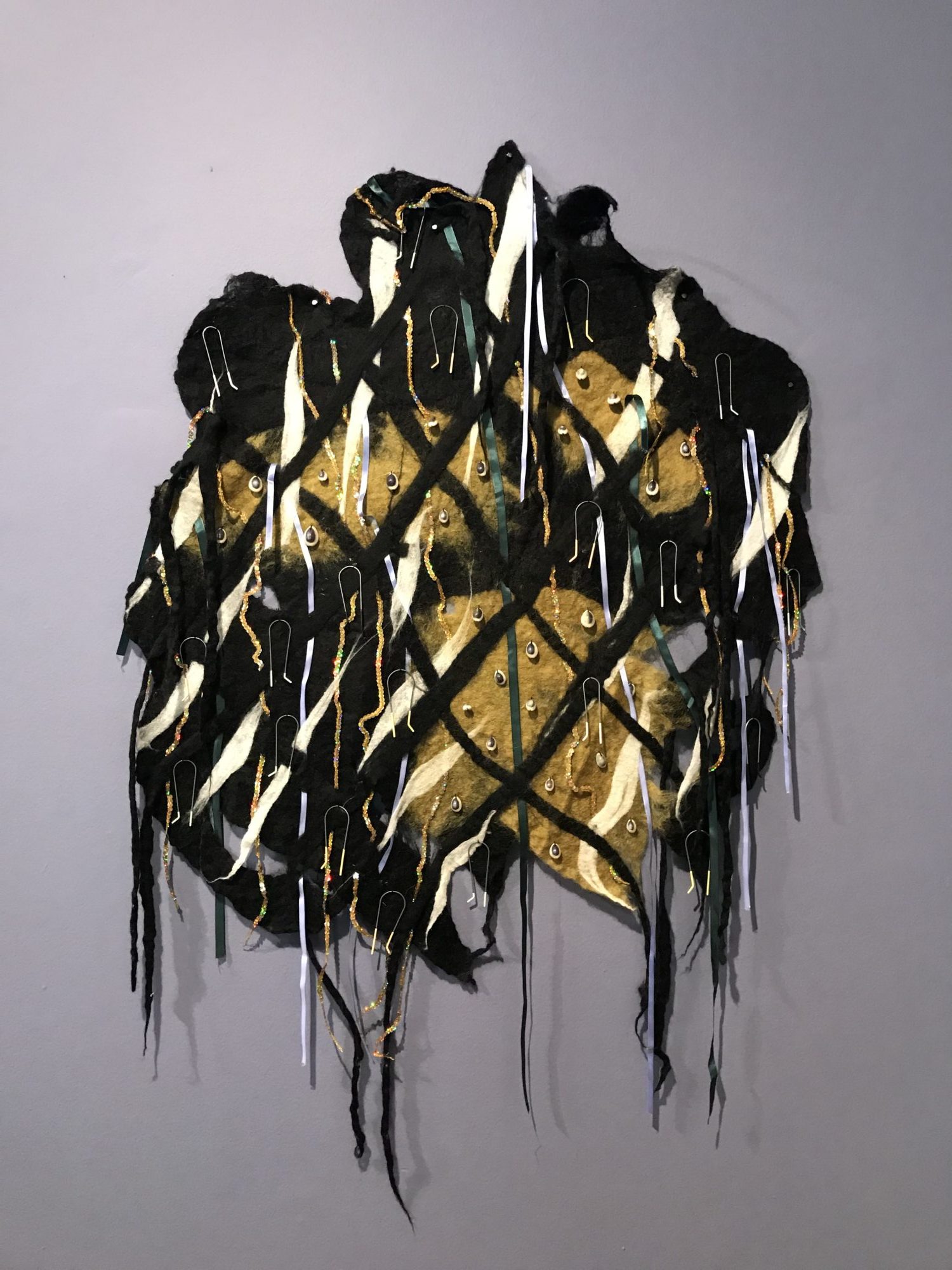 organic looking gold and black wall sculpture with hair pins and tassels hanging down against a gray wall